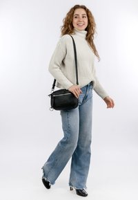Kobieta uśmiechająca się, ubrana w biały dzianinowy golf, jasnoniebieskie szerokie dżinsy, czarne botki na obcasie oraz niosąca czarną torbę na ramię.