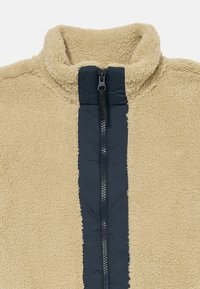 Giacca in pile beige con colletto alto, dotata di una cerniera frontale blu navy e pannello a contrasto, texture morbida e senza loghi visibili.