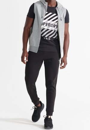 Mand iført sort t-shirt med grafik, grå ærmeløs hættetrøje med lynlås, sorte joggingsbukser og sorte sneakers, der går foran en neutral baggrund.