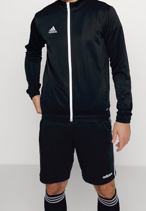 Mężczyzna noszący czarną sportową kurtkę Adidas z białym zamkiem i dopasowane czarne szorty Adidas, stojący z jedną ręką w kieszeni.