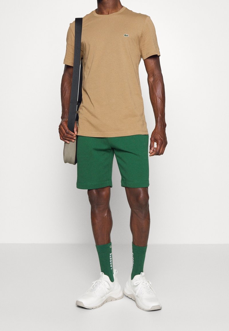 Homme portant un t-shirt beige, un short vert, des chaussettes vertes de marque, des baskets blanches, et portant un sac à bandoulière gris avec une sangle noire.