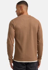 Braunes geripptes Langarmshirt mit Rundhalsausschnitt, aus strukturiertem Stoff und mit einem Lagen-Look, getragen über einem weißen Hemd.