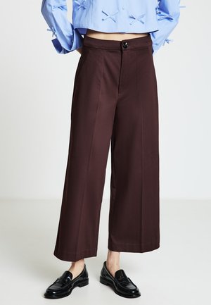 Pantaloni a gamba larga in tessuto marrone scuro dalla consistenza liscia, con chiusura frontale a bottoni e due tasche laterali. Completati da mocassini neri lucidi.