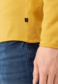 Main reposant sur le côté, portant un jean bleu et un pull jaune à manches longues avec une petite étiquette noire sur la couture latérale.