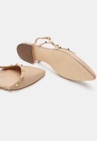 Zapatos de tacón cuadrado de charol nude con punta afilada, que presentan tachuelas en forma de pirámide doradas a lo largo de la correa para acentuar y texturizar.