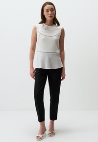 Donna in piedi che indossa una blusa bianca senza maniche con texture, pantaloni neri aderenti e sandali con tacco aperti bianchi su uno sfondo bianco semplice.
