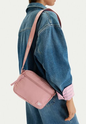 Persona con una chaqueta vaquera que lleva una pequeña bolsa bandolera rosa con una correa ancha rosa de Reebok sobre el hombro.