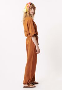 Roestkleurige jumpsuit met korte mouwen, een tailleband en taps toeloopende benen. Gemaakt van zachte stof en met geribbelde enkelaccenten met strik.