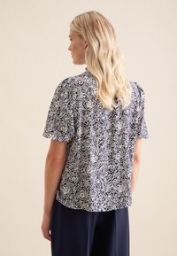 Blusa navy a maniche corte con motivo floreale bianco, tessuto leggero e vestibilità comoda. Presenta una chiusura con bottone sul retro e un orlo arrotondato.