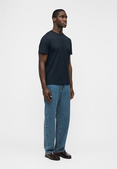 Άνθρωπος που στέκεται με το πρόσωπο προς τα εμπρός, φοράει απλό μαύρο μπλουζάκι με κοντά μανίκια, μπλε τζιν και καφέ δερμάτινα παπούτσια σε λευκό φόντο.