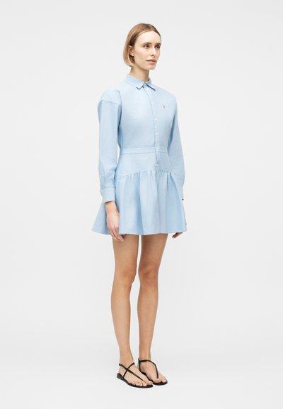 Γυναίκα που στέκεται, φοράει ένα ανοιχτό μπλε φόρεμα με μακρύ μανίκι και γιακά, και μαύρα σανδάλια με λουράκια σε λευκό φόντο.