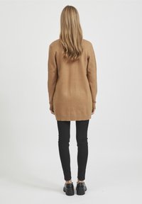 Larga chaqueta de punto marrón con dobladillo acanalado, llevada sobre pantalones ajustados negros y zapatos negros, vista desde la parte de atrás. Textura suave y corte relajado.