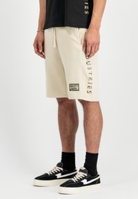 Beige bomuldsshorts med justerbar snøre, sidelommer og et logo-patch på benet. Båret med sorte sneakers og strømper.