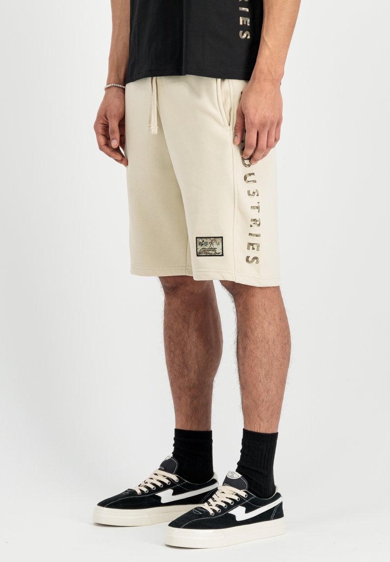 Beige bomuldsshorts med justerbar snøre, sidelommer og et logo-patch på benet. Båret med sorte sneakers og strømper.