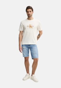 Camiseta de algodón blanca con un logo gráfico, pantalones cortos de mezclilla azul con bordes deshilachados y zapatillas blancas. El modelo está de pie frente a un fondo liso.