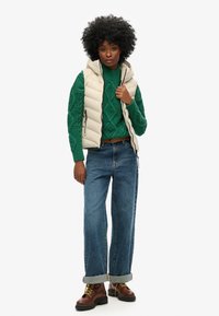 Un gilet matelassé beige sur un pull en maille torsadée verte, associé à un jean ample bleu clair et des bottes à lacets marron avec des lacets jaunes.