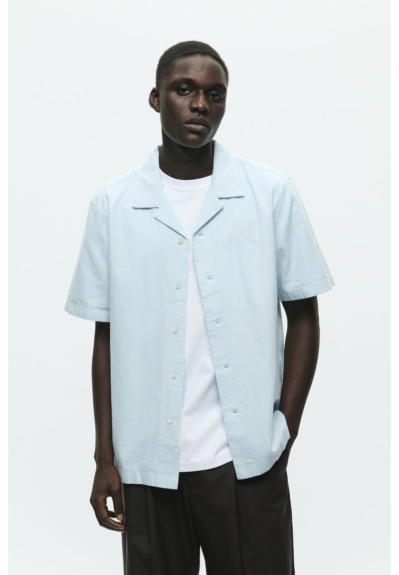 Chemise à manches courtes bleu clair avec un col camp et une fermeture à boutons. Portée ouverte sur un t-shirt blanc, avec une texture subtile et une coupe décontractée.