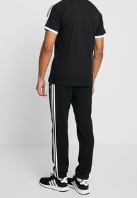 Svart sporttröja med vita detaljer, i kombination med svarta byxor med tre vita ränder längs sidorna. Skorna är svarta och vita.