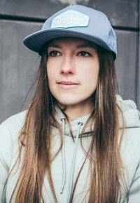 Light blue cap with a white patch featuring text; grey hoodie with drawstrings; long, straight brown hair; neutral background.
