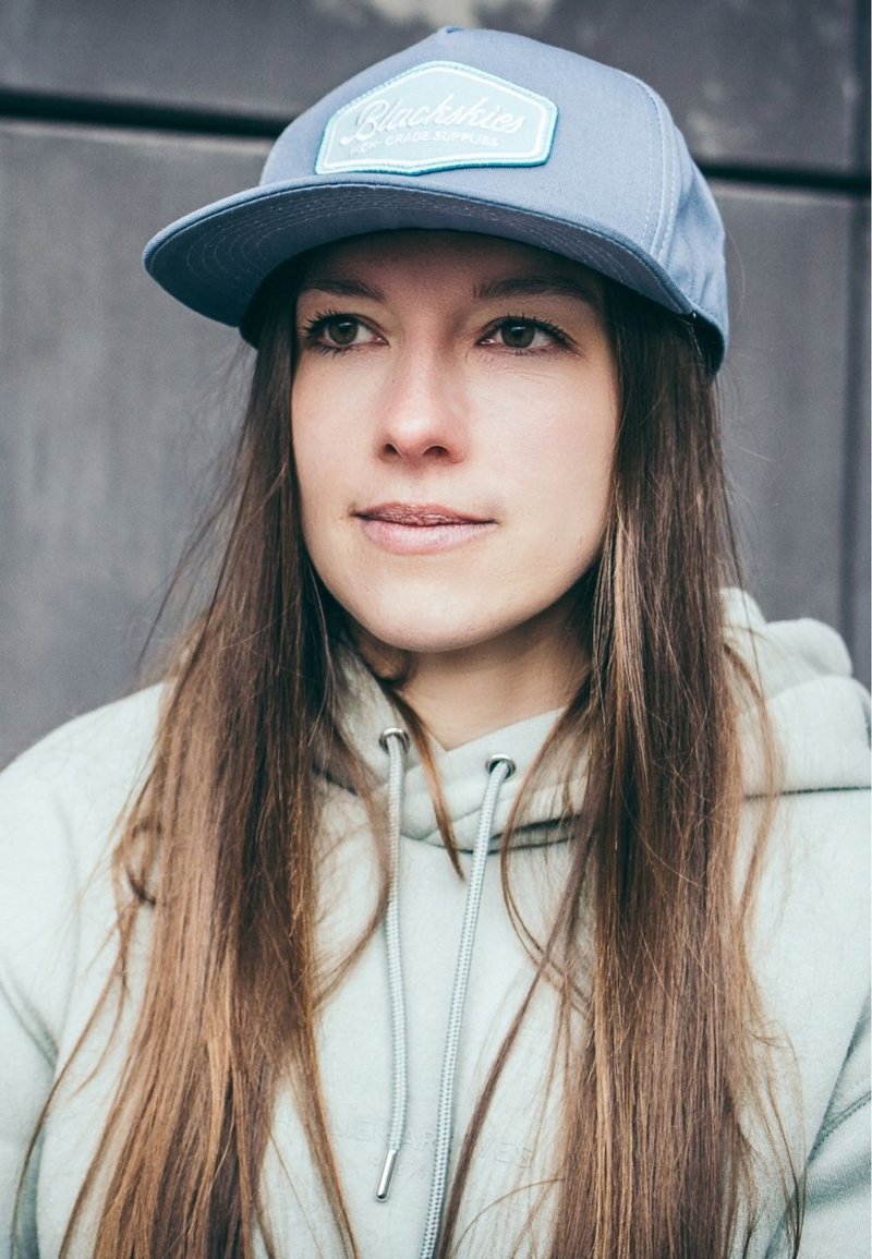 Light blue cap with a white patch featuring text; grey hoodie with drawstrings; long, straight brown hair; neutral background.