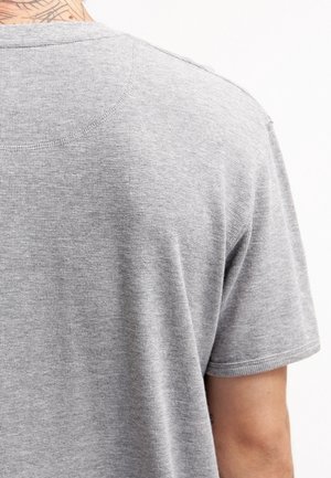 Homme portant une chemise à manches courtes gris clair, montrant le haut du dos et une partie du bras droit sur un fond blanc.