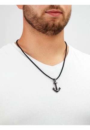 Schwarze geflochtene Halskette mit einem Ankeranhänger in metallischem Finish, getragen über einem weißen V-Ausschnitt-Shirt, das Textur und Detail betont.