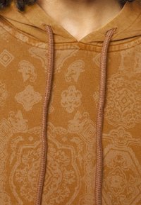 Primer plano de una persona que lleva una sudadera con capucha marrón con sutiles patrones de paisley y geométricos, y cordones a juego en marrón.