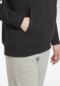 Felpa nera con polsini e bordo a coste, dotata di una tasca frontale. Abbinata a pantaloni della tuta grigi chiari, che mostrano una texture morbida.