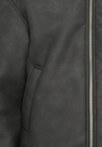 Veste en simili cuir noir avec une texture lisse, fermeture éclair et poche latérale du côté gauche.