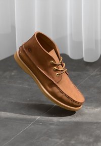 Zapato de cuero marrón con punta redondeada, dos ojetes para cordones y suela de goma. Presenta costuras a lo largo del borde y un logo en el lateral.