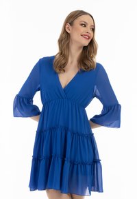 Rochie albastră, fluidă, cu decolteu adânc în V, mâneci evazate 3/4 și volane stratificate. Confecționată dintr-un material ușor, cu o textură moale.
