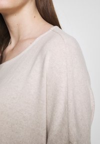 Ljus beige tröja med en mjuk, texturerad yta, med en vid halsringning och avslappnad passform. Tyget faller försiktigt över axeln.