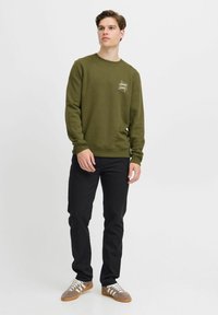 Sudadera verde oliva con cuello redondo y un logotipo sutil; combinada con jeans negros y zapatillas marrones con cordones blancos.