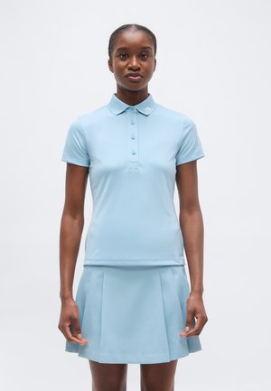 Žena stojaca čelom vpred, oblečená v svetlomodré polo tričko s krátkym rukávom a zodpovedajúcu plisovanú sukňu na jednoduchom pozadí.