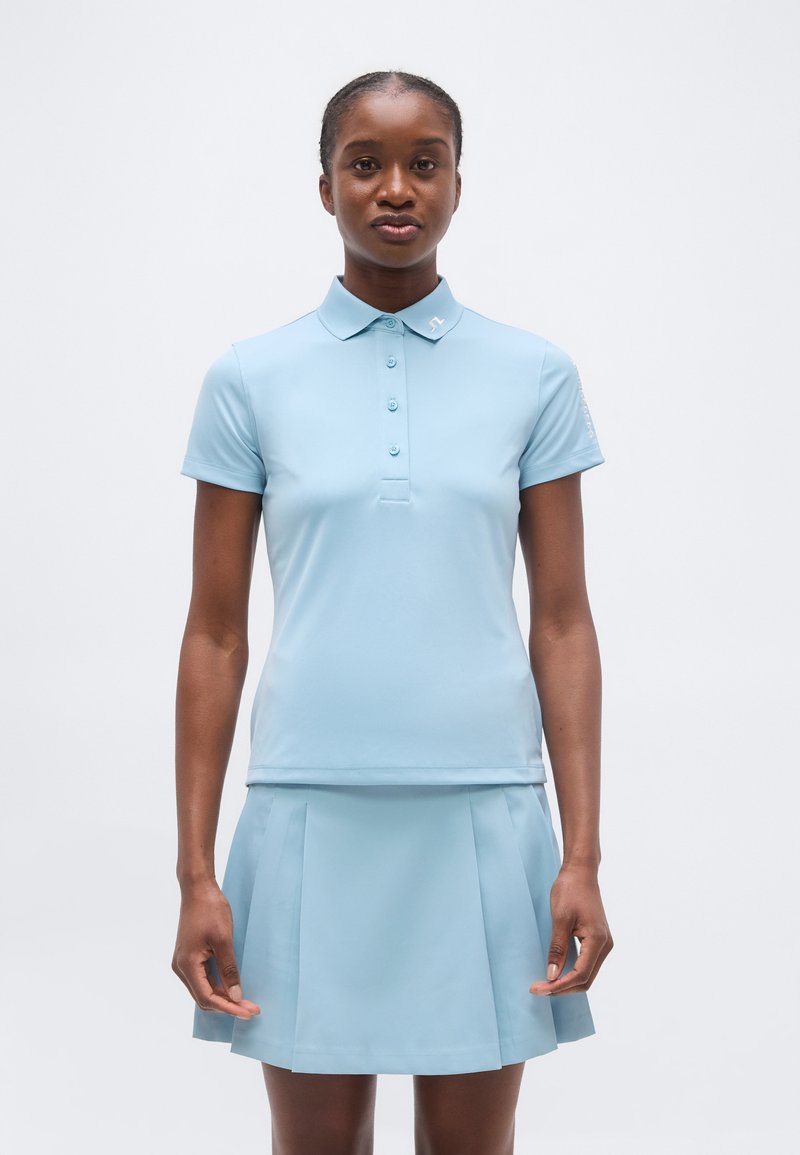 Γυναίκα που στέκεται κοιτώντας προς τα μπροστά, φορώντας ανοιχτό μπλε κοντομάνικη μπλούζα πόλο και ταιριαστό φούστα με πιέτες πάνω σε απλό φόντο.