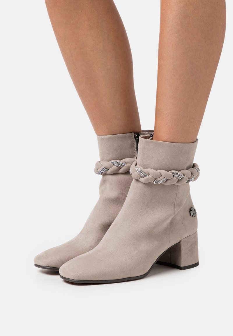 Bottines en daim beige à talons blocs, avec une bride décorative tressée ornée de strass autour des chevilles, et une fermeture éclair sur le côté intérieur.