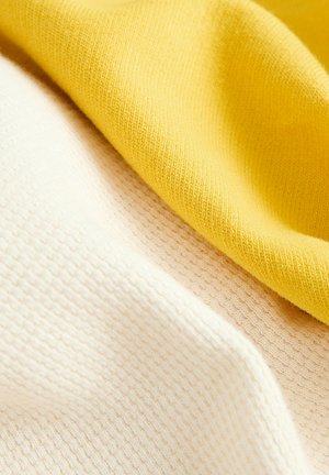 Bomullstyg i gult och krämfärgat, med subtila texturerade mönster. Den gula delen är slät medan den krämfärgade har en ribbad yta.