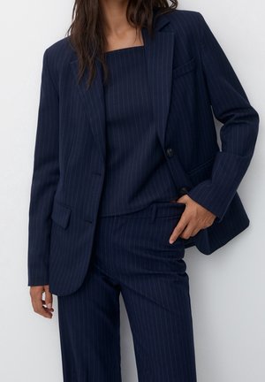 Woman wearing navy blue pinstripe suit with blazer, square-neck top, and matching trousers, hand in pocket against plain background.