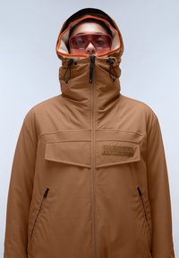 Giacca impermeabile marrone con collo alto, zip frontale, due tasche con zip e una toppa con logo. Presenta un cappuccio regolabile e un rivestimento arancione a contrasto.