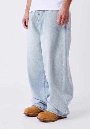 Homme portant un jean large bleu clair et des bottes en daim marron clair, debout devant un fond blanc uni.