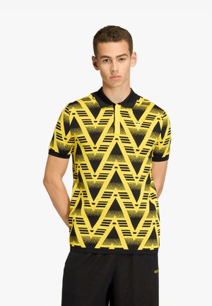Joven con una camiseta polo con patrón geométrico en amarillo y negro, con cuello negro y pantalones cortos negros, de pie con las manos detrás de la espalda.
