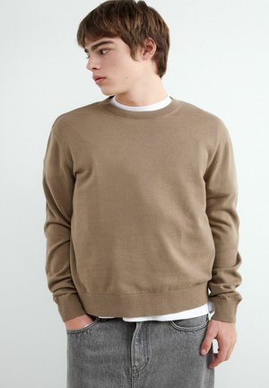 Junger Mann trägt beigen Pullover über weißem Hemd und grauen Jeans, blickt seitlich mit einer Hand in der Tasche vor schlichter Hintergrund.
