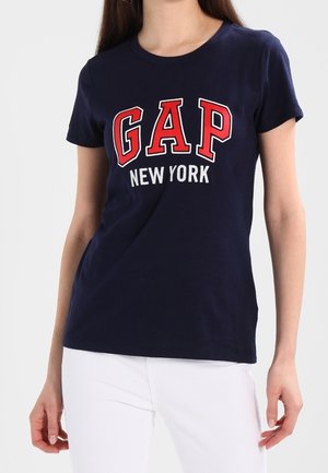 Kobieta ubrana w granatowy T-shirt z napisem „GAP New York” czerwonymi i białymi literami, w białych spodniach, stojąca na białym tle.