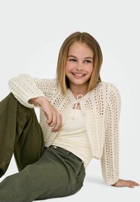 Háčkovaný krémový sveter s otvoreným vzorom, uzlový spodok, v kombinácii s ľahkým tielkom a olivovo zelenými nohavicami. Textúrovaná pletenina.