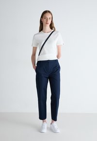 Hvit kortermet t-skjorte, marineblå bukser og hvite joggesko. Modellen har p�å seg en mørk skulderveske. Glatt stoff, skreddersydd passform, minimalistisk design.