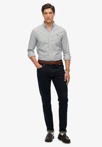 Man staat in een lichtgrijze button-up shirt met opgerolde mouwen, donkere slim jeans, een bruine gevlochten riem, donkere instappers en een horloge.