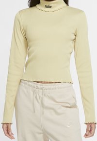 Långärmad ribbad polokrage topp i ljusgul, med en kortad design och volangkrage; svart Nike-logotyp vid nacken.
