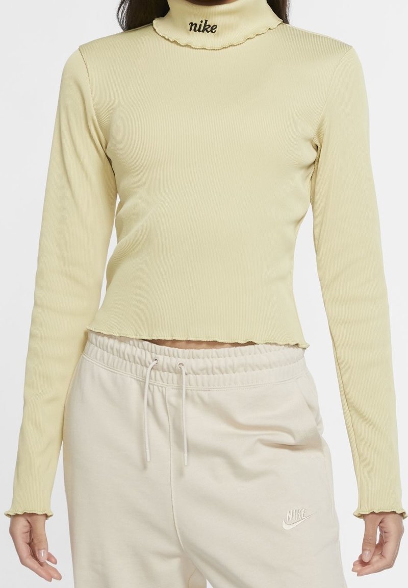 Långärmad ribbad polokrage topp i ljusgul, med en kortad design och volangkrage; svart Nike-logotyp vid nacken.