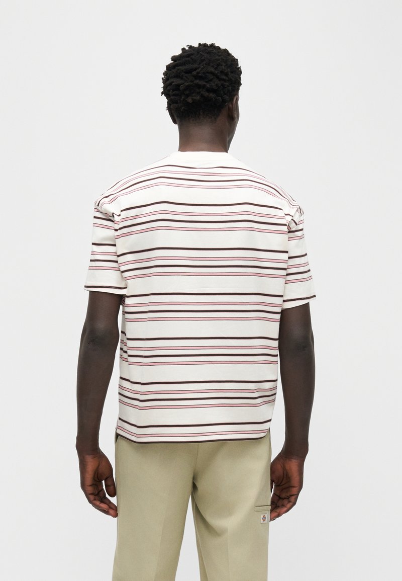 Hombre con camisa blanca de manga corta con franjas horizontales rojas y marrones, y pantalones beige, de espaldas contra un fondo liso.