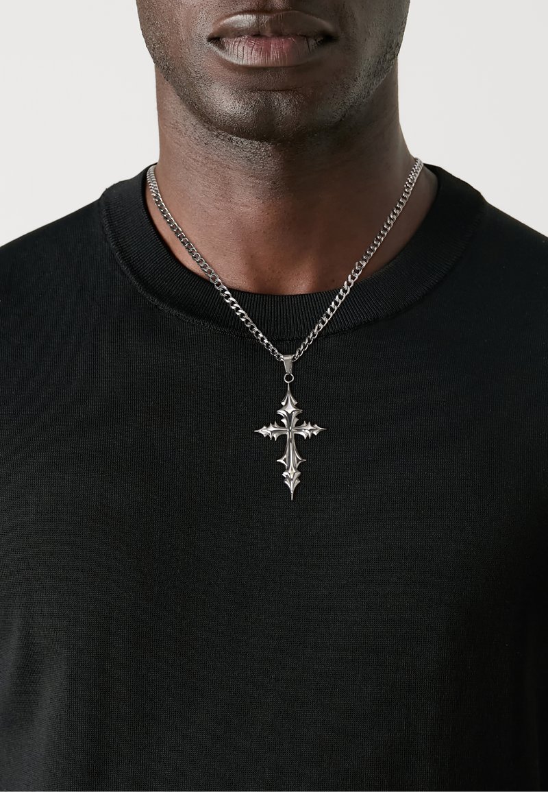 Silbernes Kreuzanhänger an einer dicken Kette, getragen über einem schwarzen strukturierten Pullover. Der Anhänger zeichnet sich durch filigrane Details und spitze Spitzen aus.